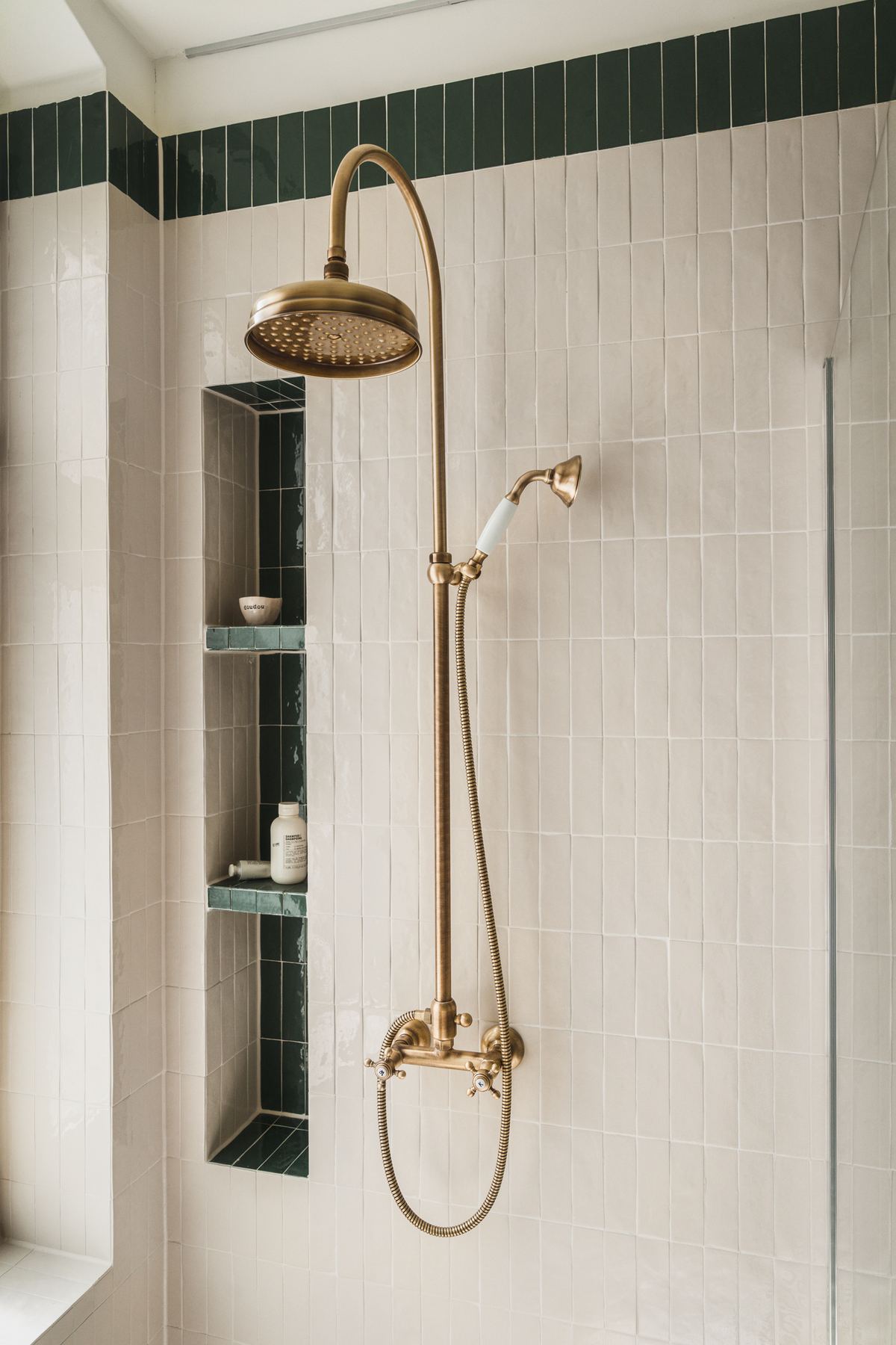 A brass rain shower on white tiled walls, with a recessed green-tiled shelf holding bottles.