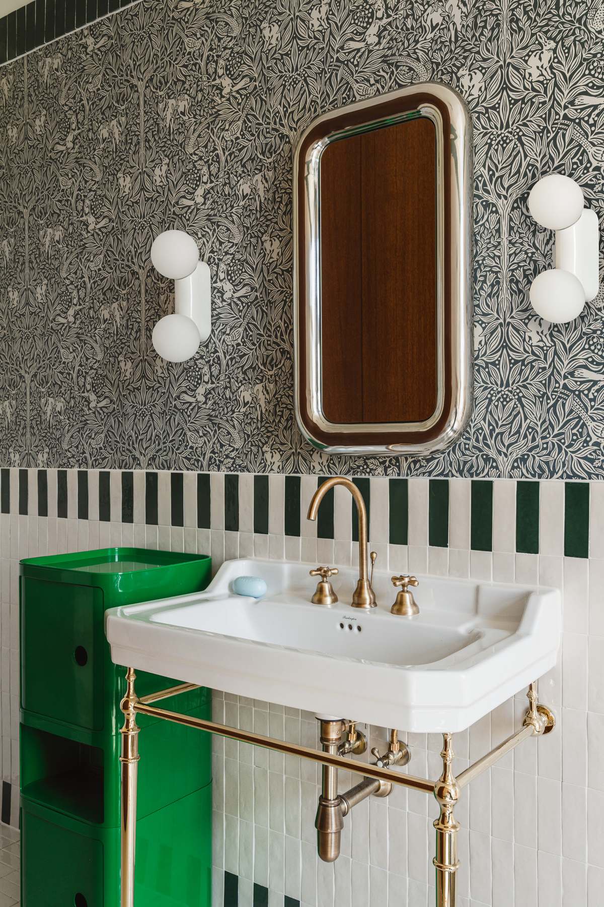 The bathroom sink on a brass stand, with green and white striped tiles below and a patterned wallpaper above.