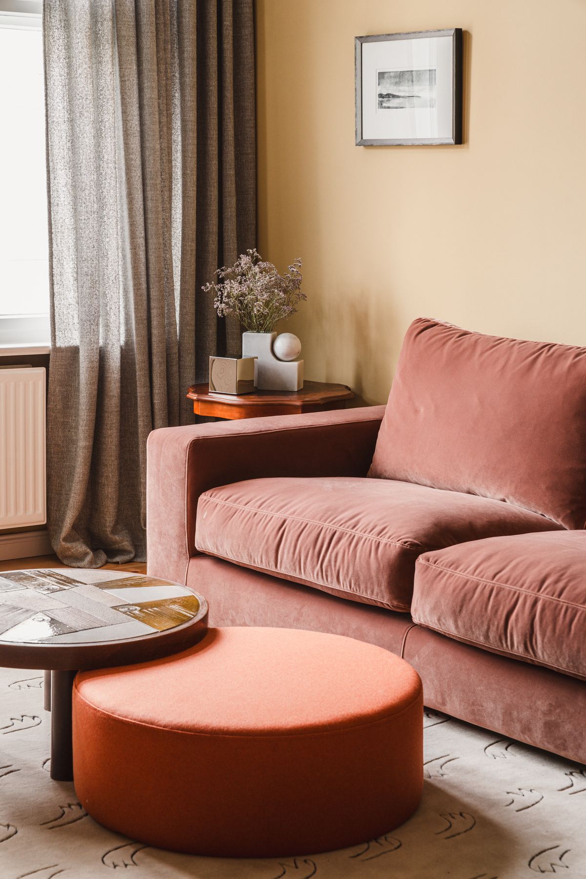 The study, with pale yellow walls, a dusty pink velvet sofa, an orange round ottoman and a curtained window.
